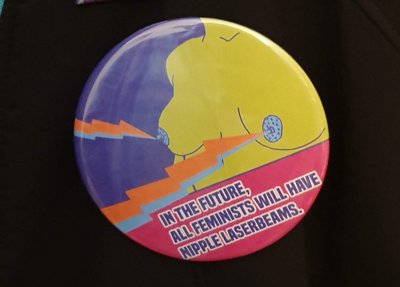 a black coat with a round badge on. The badge image is of a naked human torso with green skin and blue nipples. There are lightning bolts shooting from the breasts. The badge text reads "in the future, all feminists will have nipple laserbeams"